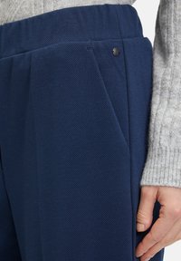 Pantaloni tailored blu navy realizzati in tessuto strutturato, con fronte plissettato, vita elastica e un piccolo dettaglio di bottone lucido.