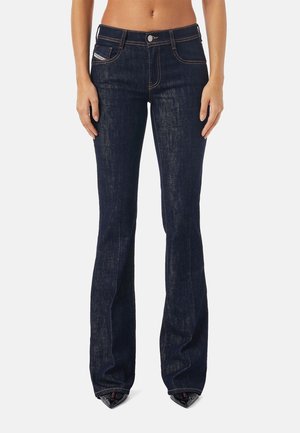 Femme portant un jean bootcut bleu foncé avec des coutures visibles et une fermeture par bouton, debout droite sur un fond uni.