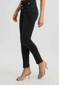 Jeans skinny neri a vita alta con una superficie liscia, dotati di un logo dorato in vita e una vestibilità aderente, abbinati a tacchi con stampa animale.