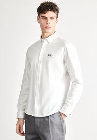 Biela košeľa s dlhým rukávom a golierom na gombíky, s malým čiernym logom „BOSS“ na ľavej strane hrudníka. Hladká bavlnená látka.