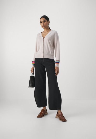 Gilet rose clair avec un décolleté en V et une fermeture à boutons. Les manches longues sont ornées d'accents festonnés multicolores. Assorti à un jean noir taille haute et des chaussures marron.