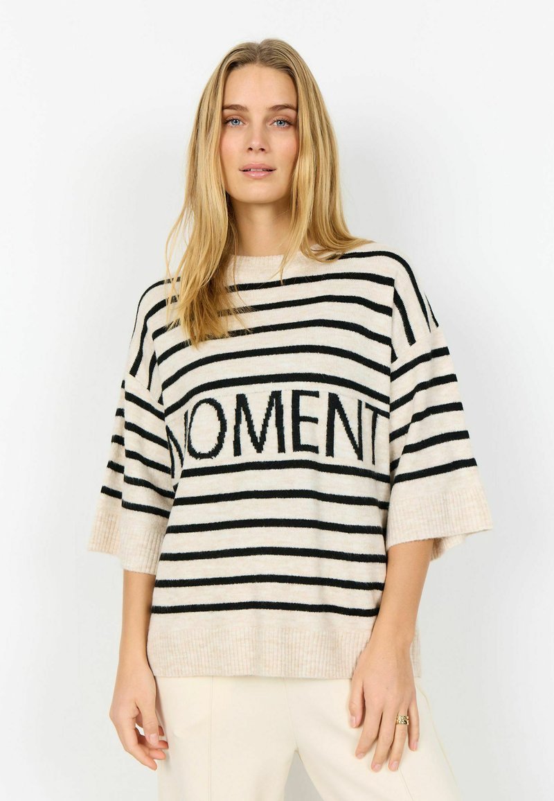 Junge Frau mit langen blonden Haaren, die einen beige-schwarz gestreiften Pullover mit dem Schriftzug "MOMENT" und cremefarbene Hosen trägt, steht vor weißem Hintergrund.