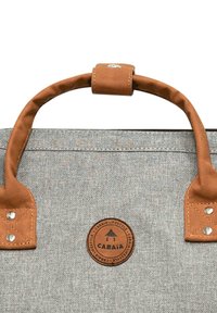 Grau gestreifte Stofftasche mit braunen Lederakzenten, einem kreisförmigen Logo-Patch und robusten genähten Griffen. Verfügt über silberne Hardware und eine strukturierte Oberfläche.