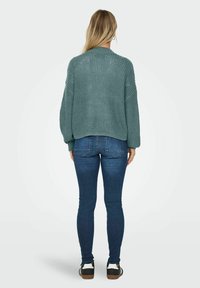 Pleteni, veliki teal pulover s nisko postavljenim šavovima ramena, uparen s tamnoplave slim-fit traperice i crno-bijele tenisice.