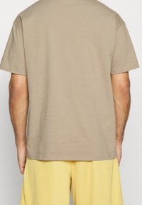 Camiseta de algodón beige con mangas cortas, con un dobladillo recto y sin marcas visibles, combinada con pantalones cortos deportivos amarillos.
