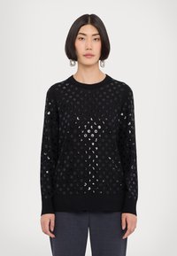 Schwarzer Pullover mit rundem Ausschnitt, langen Ärmeln und strukturierter Oberfläche mit glänzenden, kreisförmigen Mustern. Kombiniert mit grauen Hosen.