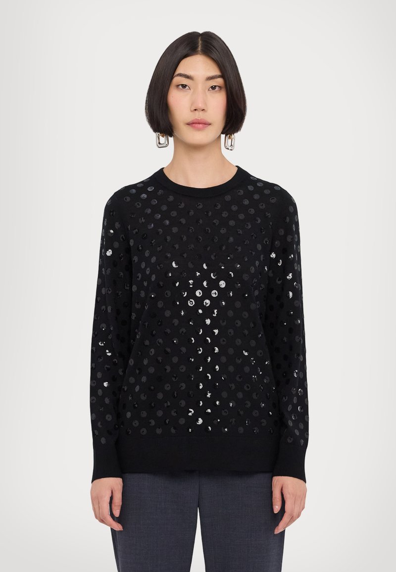 Schwarzer Pullover mit rundem Ausschnitt, langen Ärmeln und strukturierter Oberfläche mit glänzenden, kreisförmigen Mustern. Kombiniert mit grauen Hosen.
