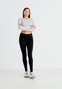 Szürke, hosszú ujjú felső fekete, testhez simuló leggingszel párosítva. A modell fehér sportcipőt visel, és szolid háttér előtt áll.