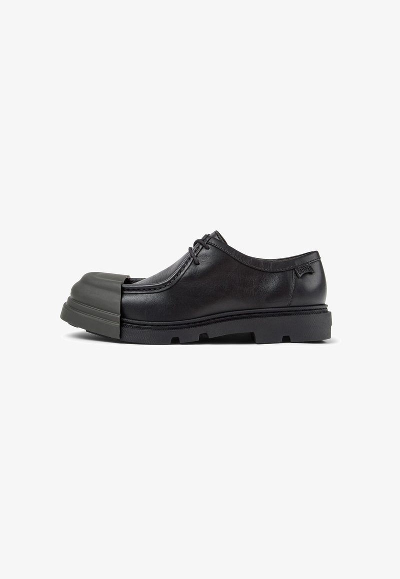 Zapato de cuero negro con punta redondeada que presenta una tapa de goma gris. Incluye un diseño de cordones y una suela texturizada para mayor tracción.