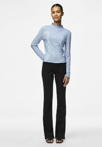 Camicia azzurro chiaro a maniche lunghe con colletto e orlo a volant, abbinata a pantaloni neri a palazzo e sandali con punta aperta. Tessuto morbido.