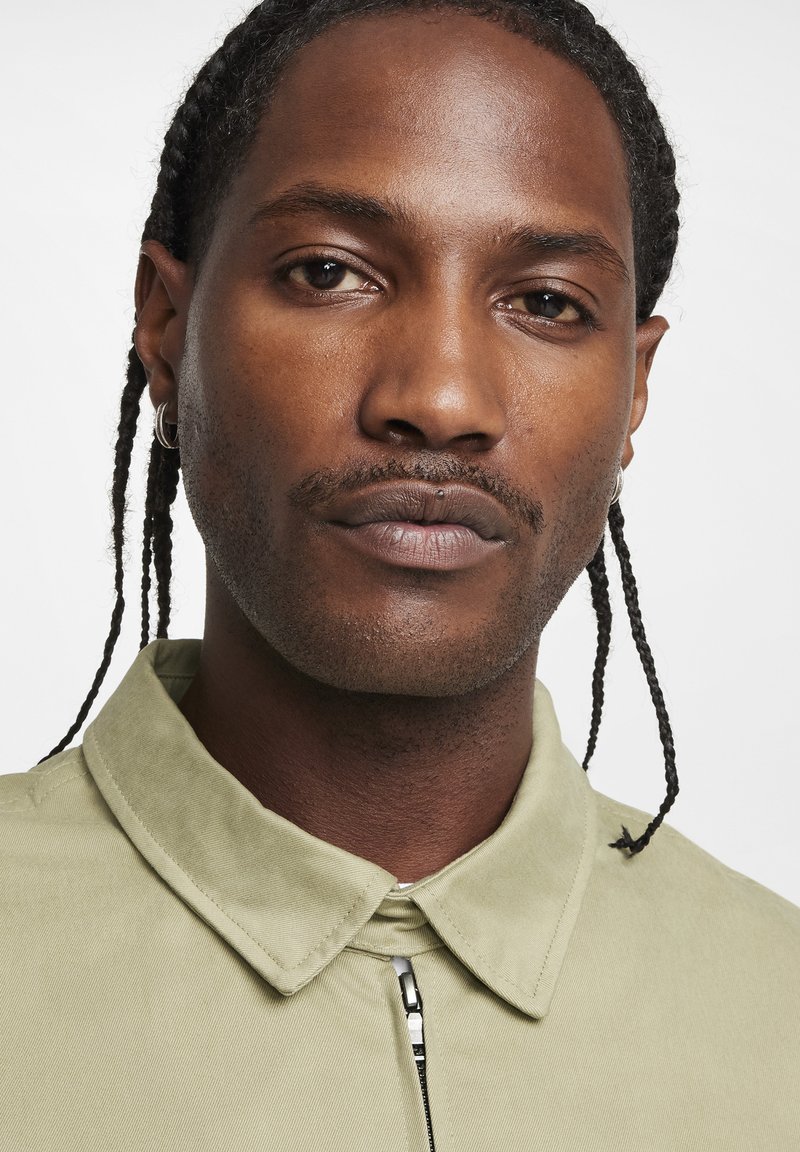 Chemise col rond vert clair avec fermeture éclair à l'avant, confectionnée dans un tissu lisse au motif subtil. Portée par une personne ayant des cheveux tressés.