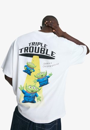 Hvid bomulds t-shirt med et grafisk print af tre tegneseriealiens i blå outfits, med teksten "TRIPLE TROUBLE" og "Rummet er vores legeplads."