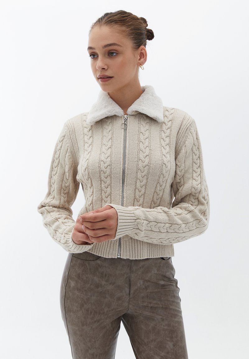 Beige kaapuknitted kardigan med vit faux fur-krage och silverfärgad dragkedja. Ribbstickade ärmslut och nederdel, med strukturerade mönster över hela plagget.