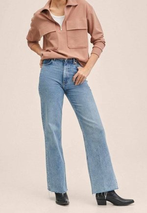 Kvinde iført en støvet lyserød sweatjakke med lynlås og store lommer, lyseblå high-waist flared jeans og sorte spidse støvler.