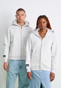 Sweatshirts zippés gris en matière douce, dotés de poches kangourou et de poignets côtelés. Les modèles portent des jeans en denim ; l'un d'eux a un accent avec une étiquette noire.