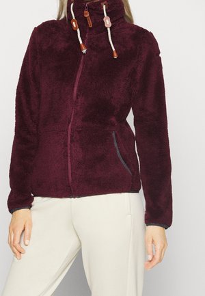 Vrouw met een bordeauxrode fleecejas met ritssluiting, hoge kraag en trekkoorden, gecombineerd met lichtbeige broeken.