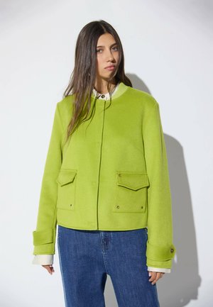 Mujer con el pelo largo y castaño que lleva una chaqueta verde brillante sobre una camisa blanca y vaqueros azules, de pie frente a un fondo blanco liso.