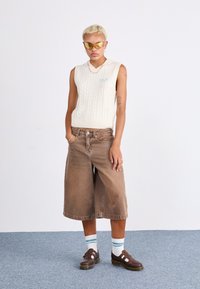 Strikket kremfarget ermela genser med kabelmønster, paret med vide brune denimshorts. Brune sko med hvite detaljer og stripete sokker.