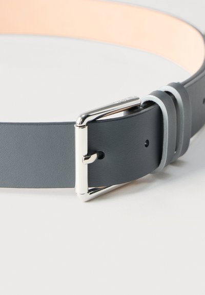 Σκούρα γκρι δερμάτινη ζώνη με ασημένια μεταλλική αγκράφα και δύο θηλιές πάνω σε λευκή επιφάνεια, εμφανιζόμενη κοντινά και μερικώς τυλιγμένη.