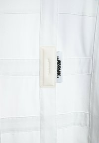 Tela blanca con paneles cosidos, con una etiqueta suave y texturizada y una etiqueta de logo negra. El diseño es minimalista y moderno.