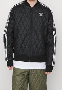 Svart vadderad jacka med dragkedja, prydd med vita tre-randiga detaljer på ärmarna och en liten logotyp på bröstet. Mjuk, vadderad yta.