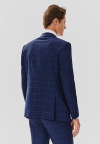 Granatowa marynarka w kratę o strukturalnym designie, z pojedynczym wentylem z tyłu i guziczkami na mankietach, pokazana od tyłu.