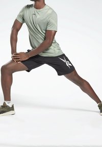 Hombre con camiseta verde y pantalones cortos negros que estira las piernas ampliamente con las manos apoyadas en una rodilla flexionada, vistiendo zapatillas olivo y calcetines blancos.