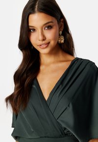 Abito di seta verde scuro con scollatura profonda, maniche drapè corte e design plissettato. Orecchini dorati e texturizzati completano il look.