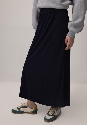 Inimene, kes kannab pikka musta seelikut, halli dressipluusi ja valgeid tossusid roheliste ja kollaste detailidega, seisab neutraalsel taustal.