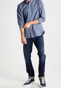 Blå skjorta med knappar och uppvikt ärm, mörka denimjeans och sneakers med vit sula och röda detaljer.