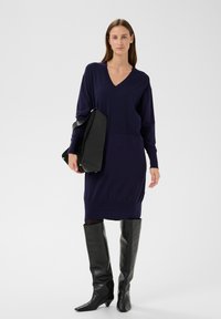 Marineblaues Strickkleid mit V-Ausschnitt, langen Ärmeln und geripptem Saum, kombiniert mit schwarzen, kniehohen Lederstiefeln mit spitzer Spitze und einer schwarzen Schultertasche.