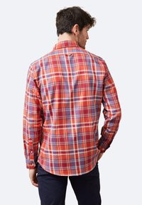 Camisa de manga larga con un patrón de cuadros en rojo, naranja y púrpura, con botones, un collar y un dobladillo curvado. La tela parece ligera.