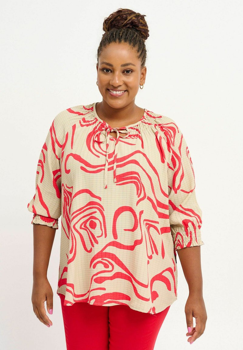 Femme aux cheveux tressés en chignon portant un chemisier beige avec des motifs abstraits rouges et un pantalon rouge, souriant devant un fond uni.