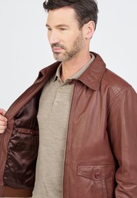 Braune Lederjacke mit einem Reißverschluss vorn, großem Kragen und einer Seiten Tasche. Innen mit glattem braunen Futter und einer zusätzlichen Tasche.