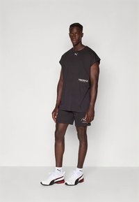 Fit man standing wearing black sleeveless Puma shirt and shorts with white Puma sneakers and socks against a plain light background.