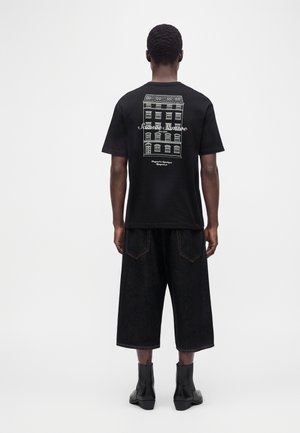 Mann trägt schwarzes T-Shirt mit weißem Gebäudegrafik und Text auf dem Rücken, lockere schwarze knielange Shorts und schwarze Stiefel, von hinten gesehen.