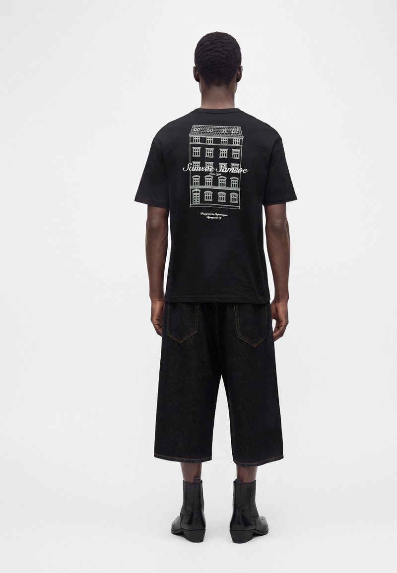 Homme portant un t-shirt noir avec un graphisme de bâtiment blanc et un texte dans le dos, un short noir ample jusqu'aux genoux, et des bottes noires, de dos.