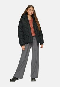 Schwarze Steppjacke mit Kapuze, die ein gestepptes Muster aufweist; getragen über einem rostfarbenen Pullover und grauen weiten Hosen. Schwarze Stiefel.