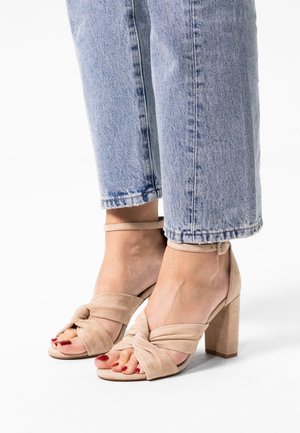 Beige heeled sandals feature a knotted front strap, smooth suede texture, and a square block heel, paired with light blue cropped jeans.
