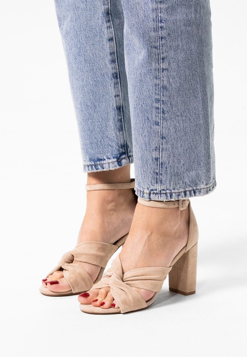 Beige hoge hakken sandalen hebben een geknoopt voorband, een zachte suede textuur en een vierkante blokhak, gecombineerd met lichtblauwe cropped jeans.