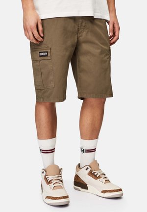 Khaki cargo shorts dengan saku samping, kain katun, potongan lurus, dan label "HONESTY" di paha kiri. Dipadukan dengan kaus kaki putih dan sepatu sneakers.