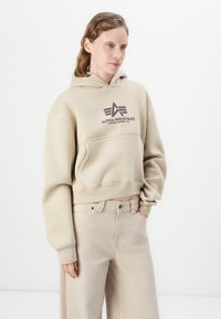 Donna che indossa una felpa beige di Alpha Industries e pantaloni a vita alta beige chiaro, con una mano in tasca, in piedi contro uno sfondo bianco puro.