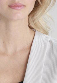 Primo piano di un colletto di blazer in tessuto a coste di un grigio chiaro, che rivela un tono di pelle levigato e lentiggini sottili su un collo. Capelli ondulati e fluenti visibili.