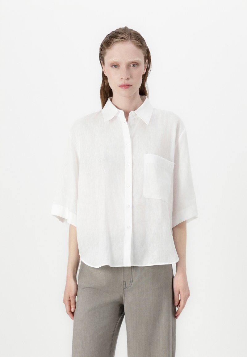 White short-sleeve shirt made of lightweight fabric. Features a button front, collar, and single chest pocket. Clean, minimal design.
