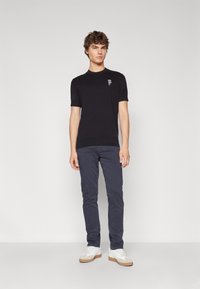 Maglietta nera a maniche corte con un piccolo design ricamato, abbinata a pantaloni blu navy e sneakers bianche con dettagli beige.