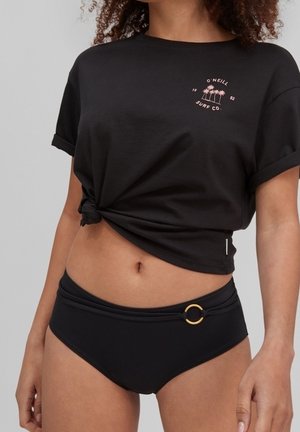 Kobieta ubrana w czarny, wiązany na supeł t-shirt z logo palmy oraz czarne bikini z detalem w postaci złotego pierścienia na lewym boku bioder.