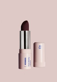 Labial de Korres True Velvety en un tubo rosa, con una tapa plateada metálica. Punta de labial burdeos profundo, con un acabado texturizado.