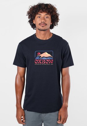 Homme aux cheveux bouclés portant un T-shirt noir avec un graphique de montagne et le mot "OXBOW" en lettres rouges et roses.
