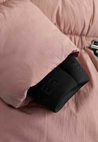 Manica di giacca a vento rosa con polsino interno nero che mostra lettere parzialmente in rilievo, con un fermaglio in metallo e dettagli di cuciture trapuntate.