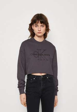 Kort grå collegegenser laget av bomull, med en stor svart "Calvin Klein Jeans" logo på brystet og ribbestrikkede mansjetter.